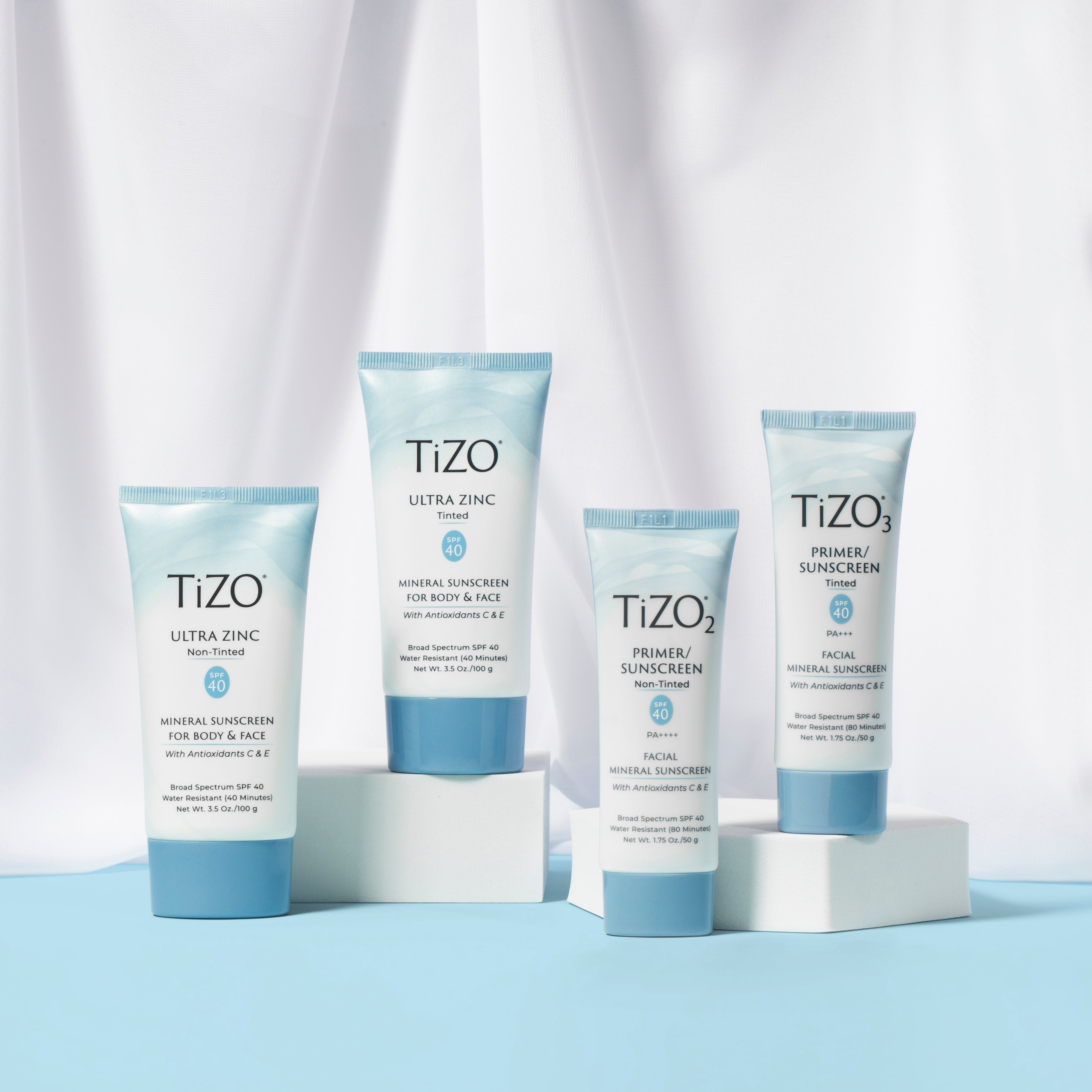 A collection of TiZo sunscreen bottles displayed on a light blue surface and white platform with a white curtain background