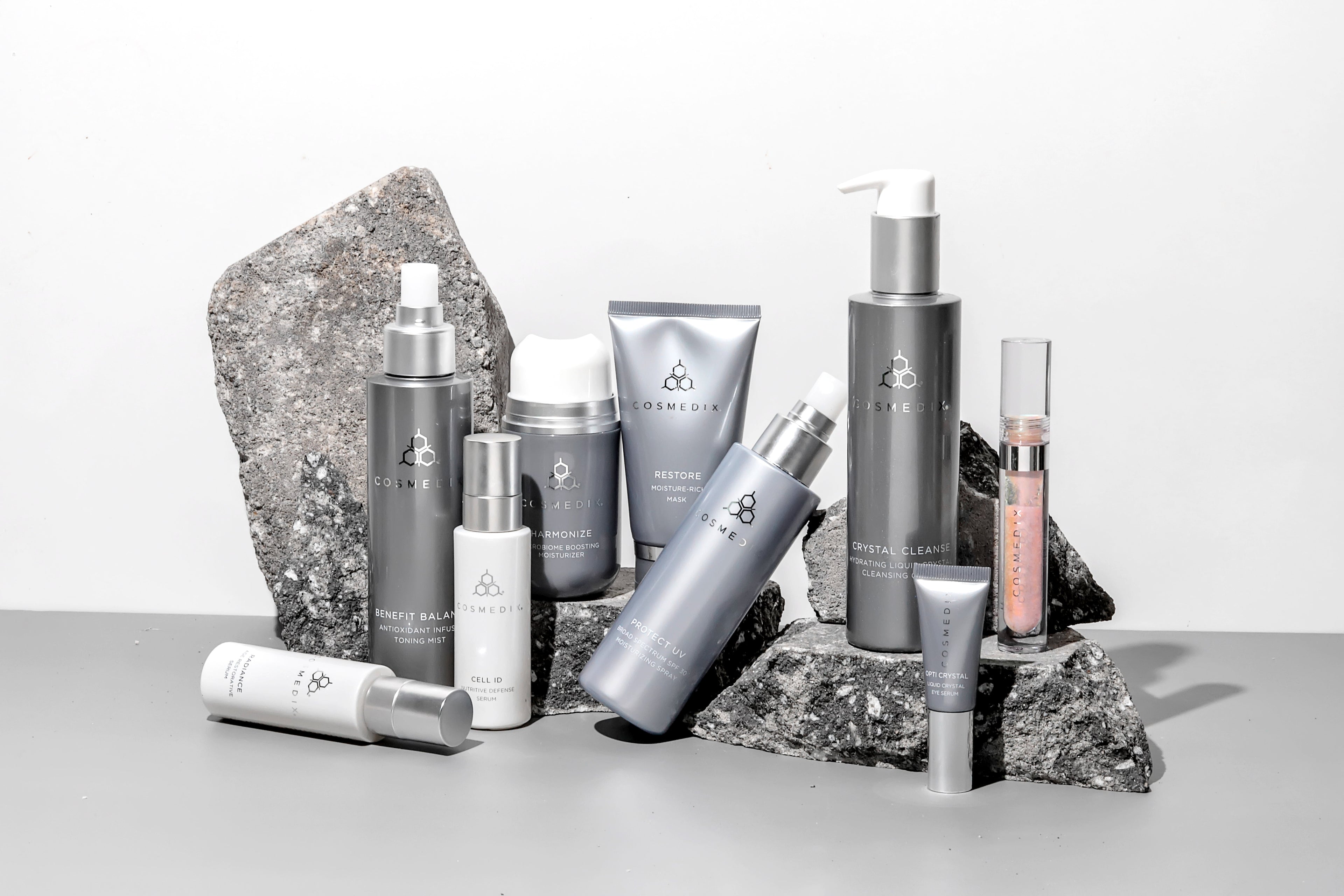 A collection of Cosmedix products displayed with grey rocks on a grey surface with a white background
