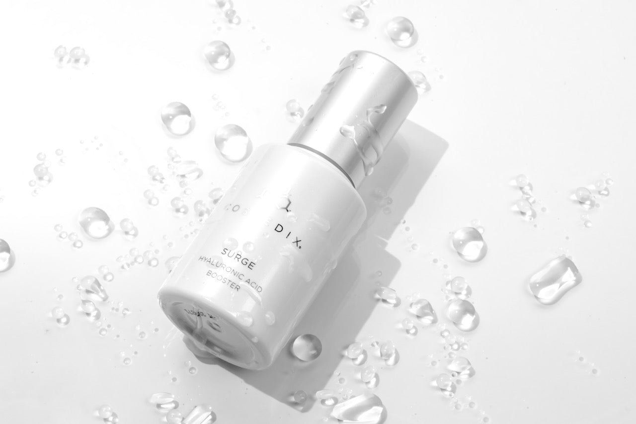 A cosmedix serum with water droplets on a white gradient background