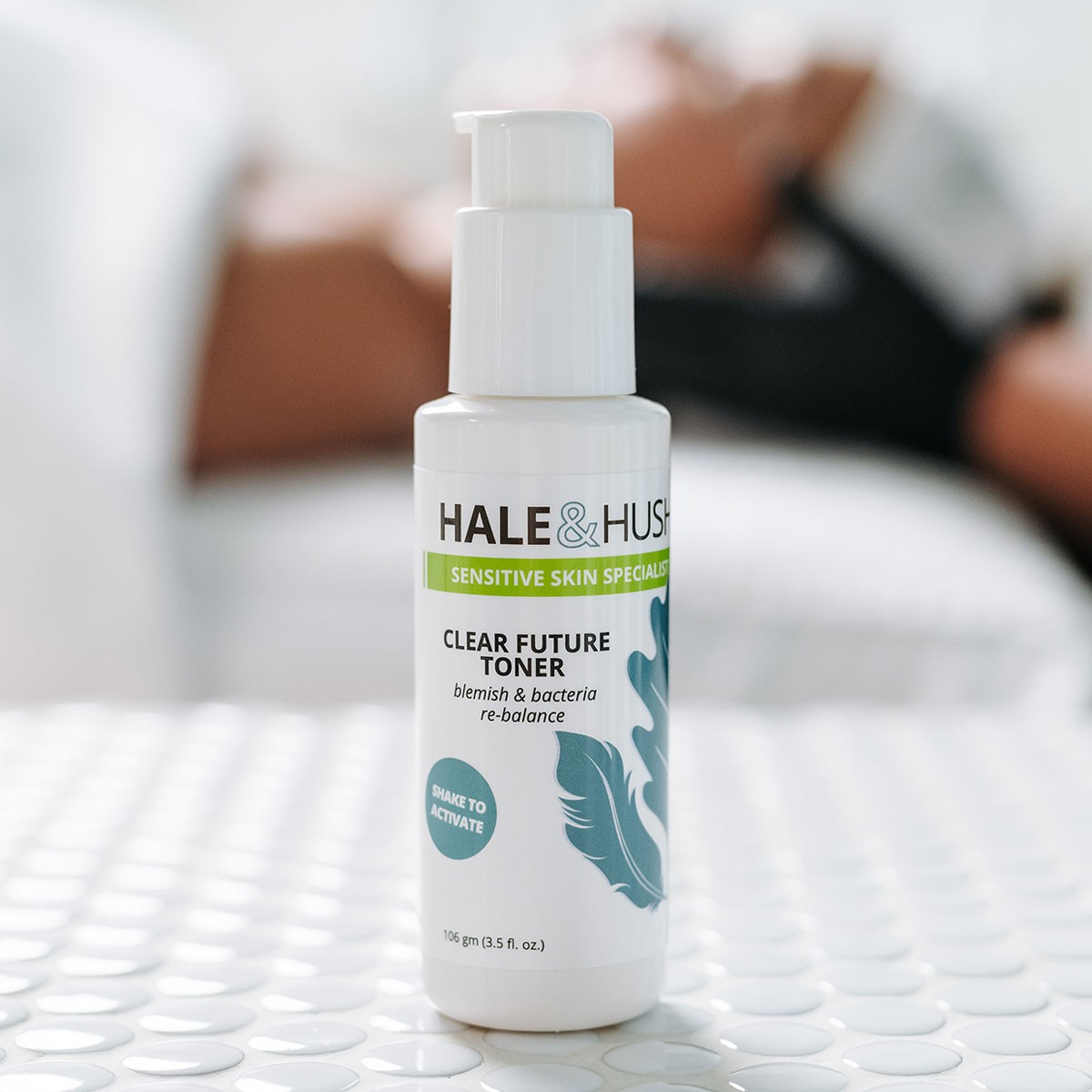 Hale & Hush's Clear Future Toner white product bottle standing on white spa bed with a model receiving a facial in the background