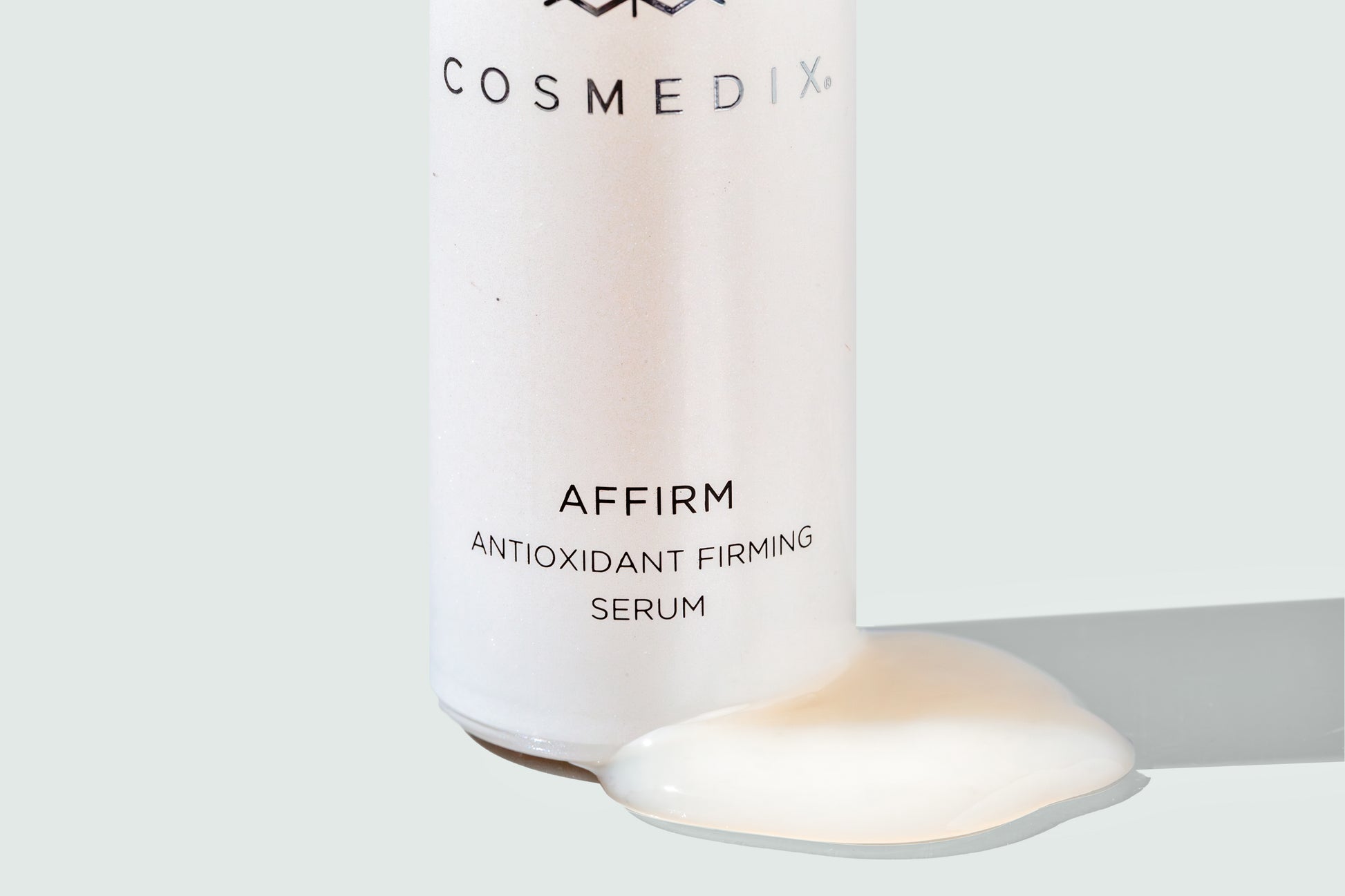 Zoomed in view of COSMEDIX's Affirm Antioxidant Firming Serum product bottle with cream colored product swatch against grey background