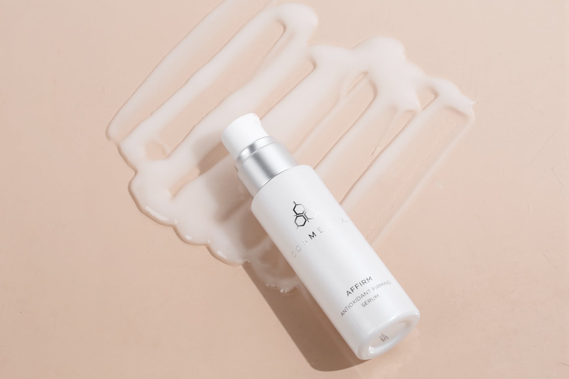 COSMEDIX's Affirm Antioxidant Firming Serum white colored product bottle laying with cream-colored product swatches against beige background