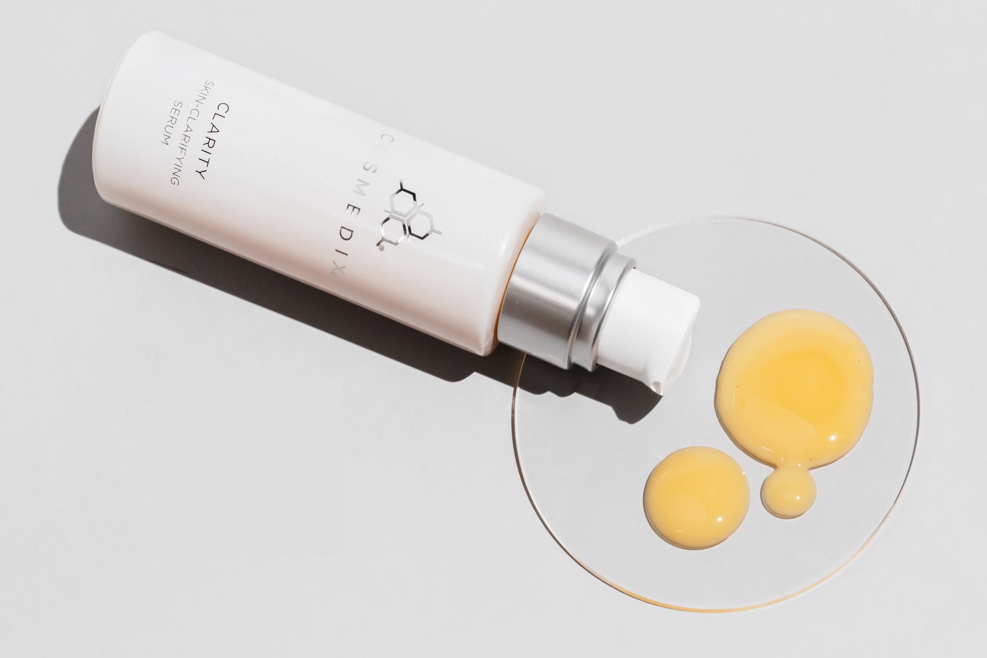 COSMEDIX's Clarity Serum product bottle laying on side with peach colored product droplets on a petri dish, against a light grey background