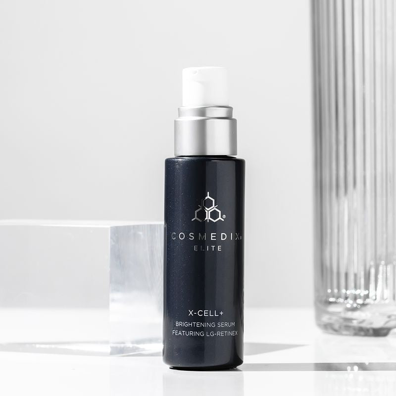 COSMEDIX's X-Cell+ Brightening Serum dark grey product bottle with white dispenser standing beside a glass cube and glass vase against light grey background