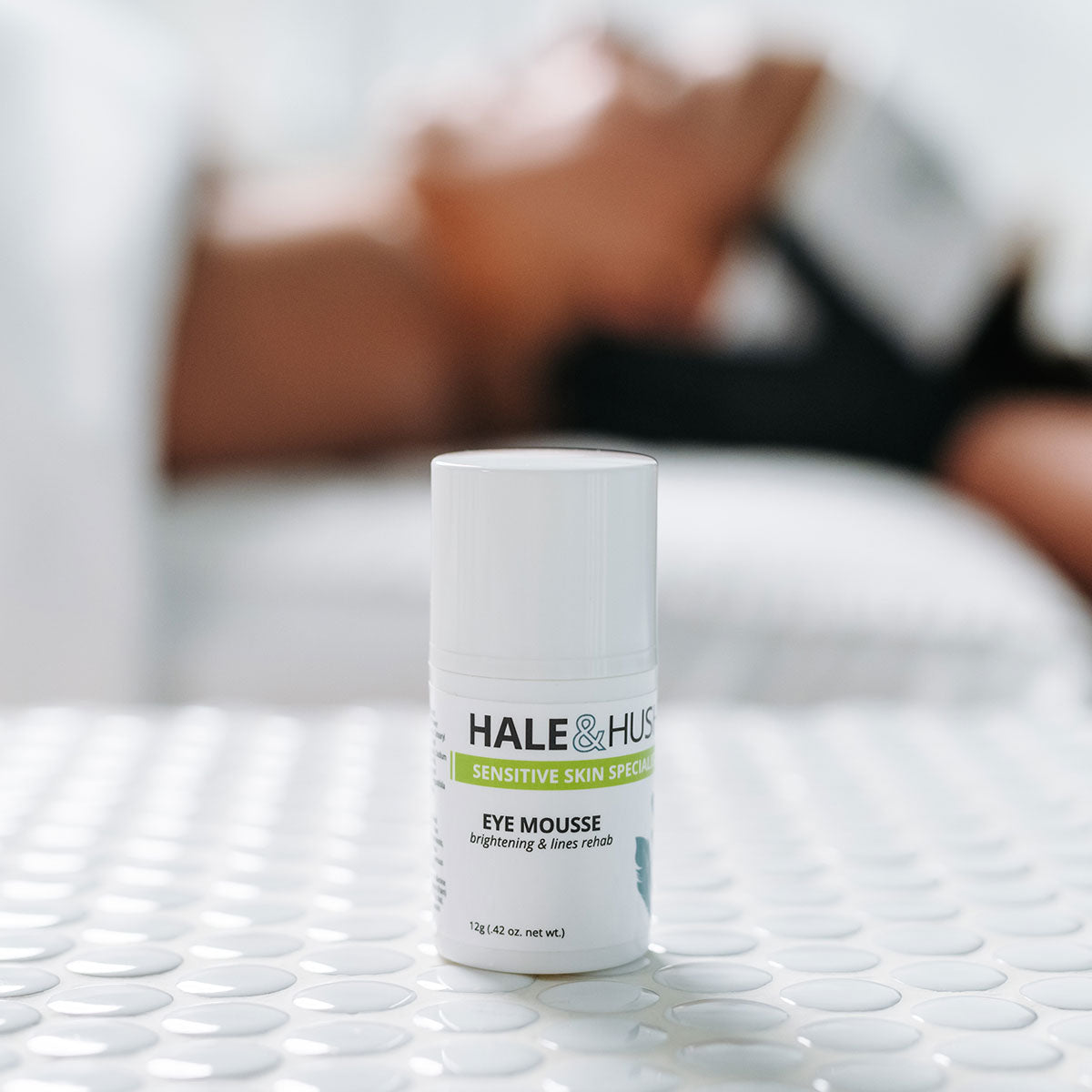 Hale & Hush's Eye Mouse white product bottle standing on white spa bed with a model receiving a facial in the background