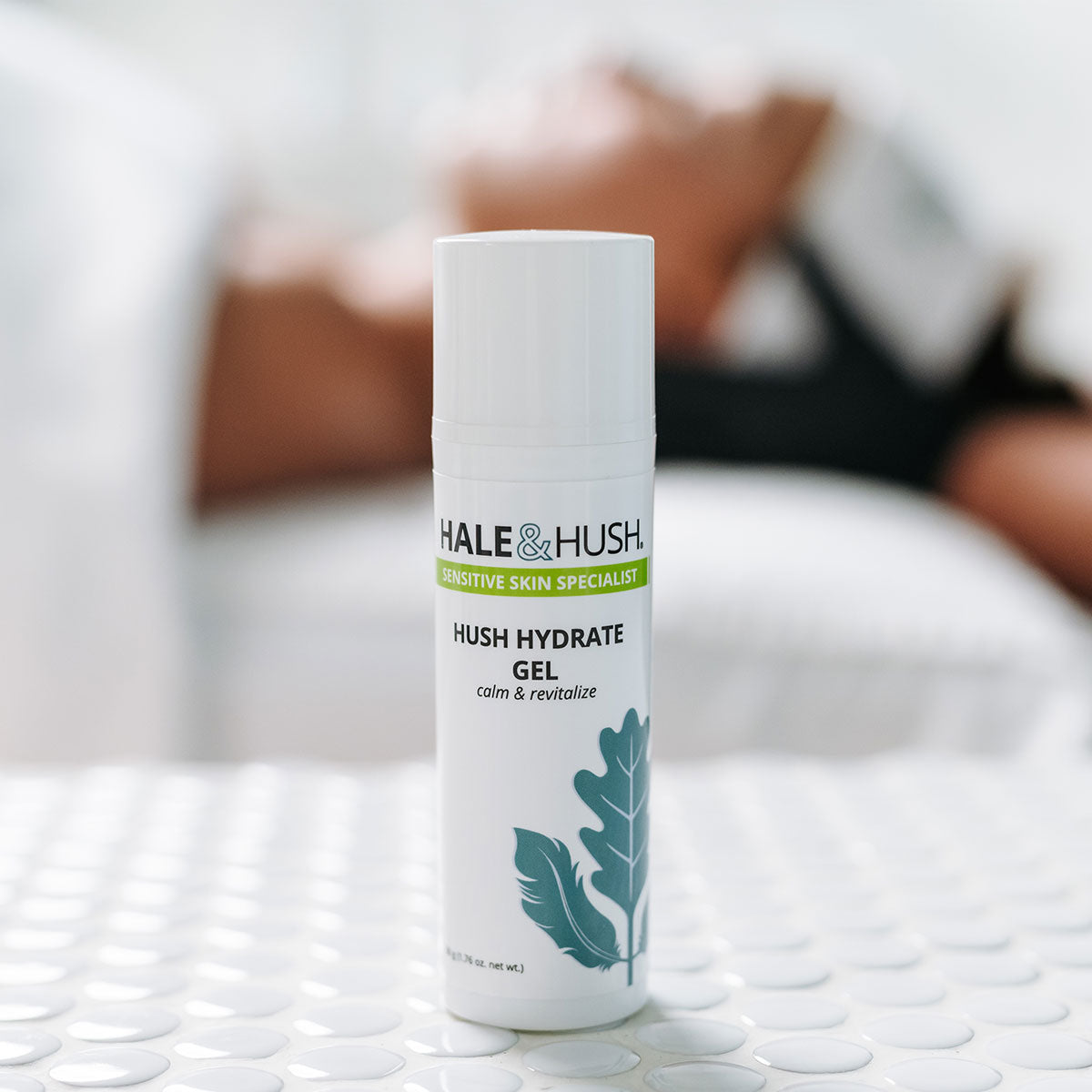Hale & Hush's Hush Hydrate Gel white product bottle standing on white spa bed with a model receiving a facial in the background