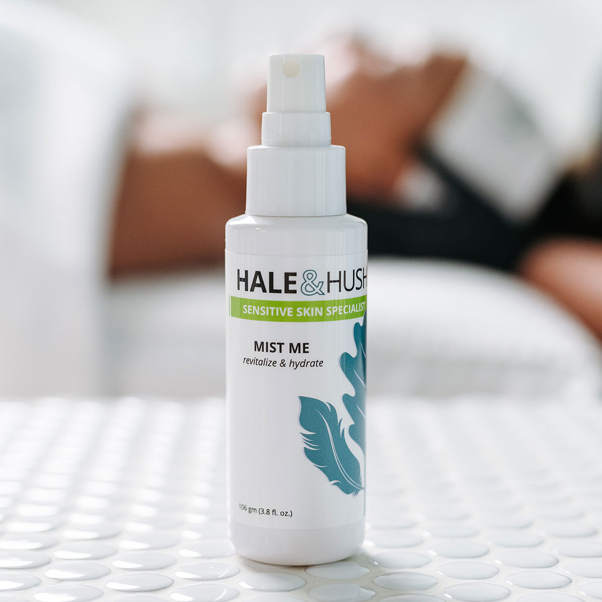 Hale & Hush's Mist Me white product bottle standing on white spa bed with a model receiving a facial in the background