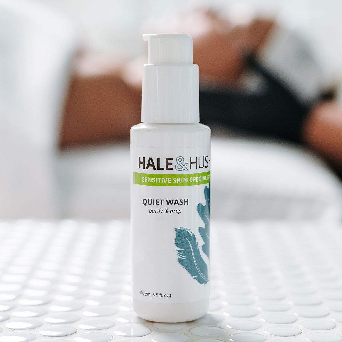 Hale & Hush's Quiet Wash white product bottle standing on white spa bed with a model receiving a facial in the background