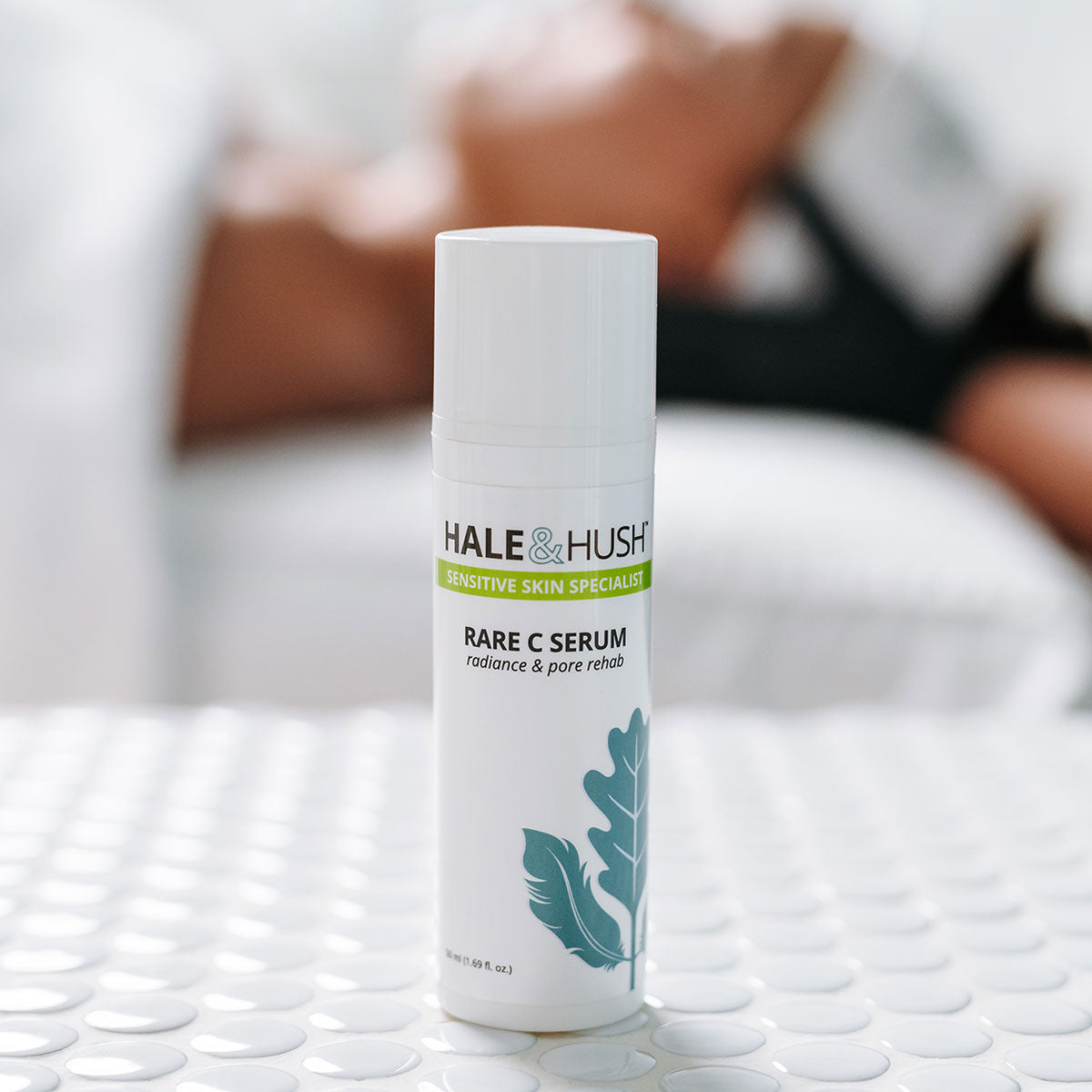 Hale & Hush's Rare C Serum white product bottle standing on white spa bed with a model receiving a facial in the background