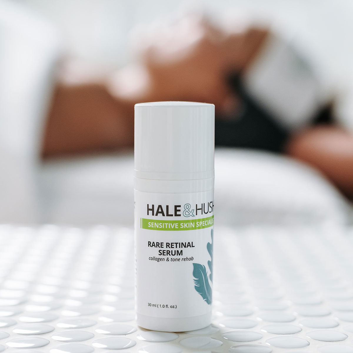 Hale & Hush's Rare Retinal Serum white product bottle standing on white spa bed with a model receiving a facial in the background