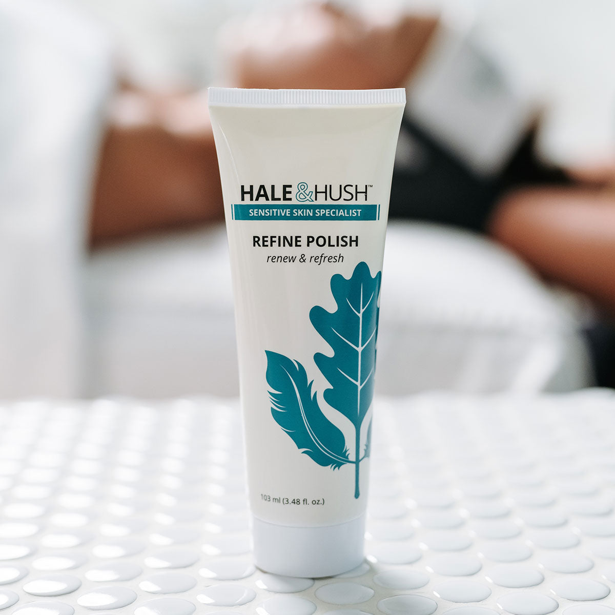 Hale & Hush's Refine Polish white product tube standing on white spa bed with a model receiving a facial in the background