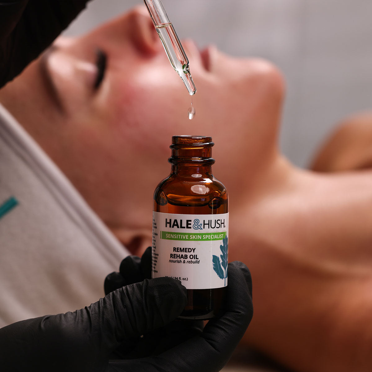 Hale & Hush's Remedy Rehab Oil bronze product bottle with dropper dispensing clear liquid with a model receiving a facial in the background