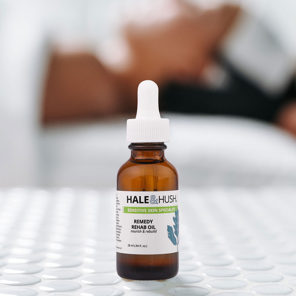 Hale & Hush's Remedy Rehab Oil bronze product bottle with dropper standing on white spa bed with a model receiving a facial in the background