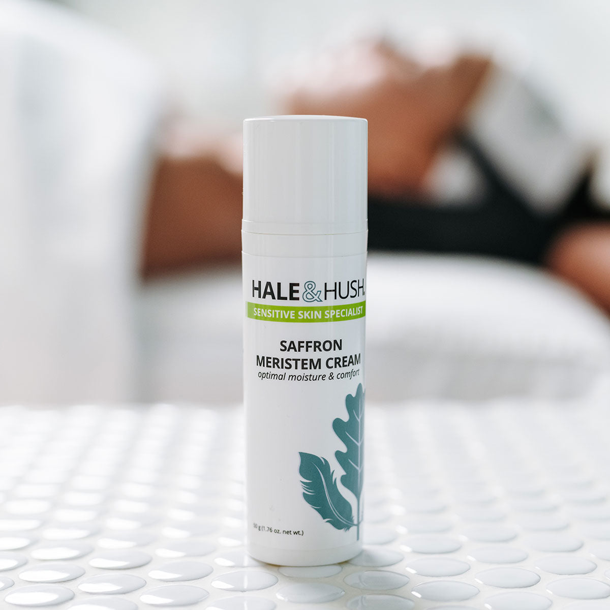 Hale & Hush's Saffron Meristem Cream white product bottle standing on white spa bed with a model receiving a facial in the background