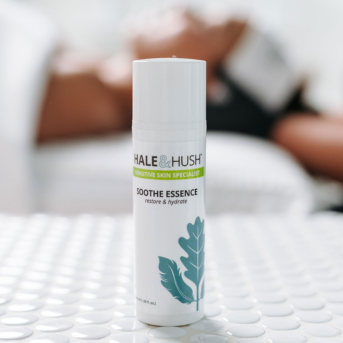 Hale & Hush's Soothe Essense white product bottle standing on white spa bed with a model receiving a facial in the background