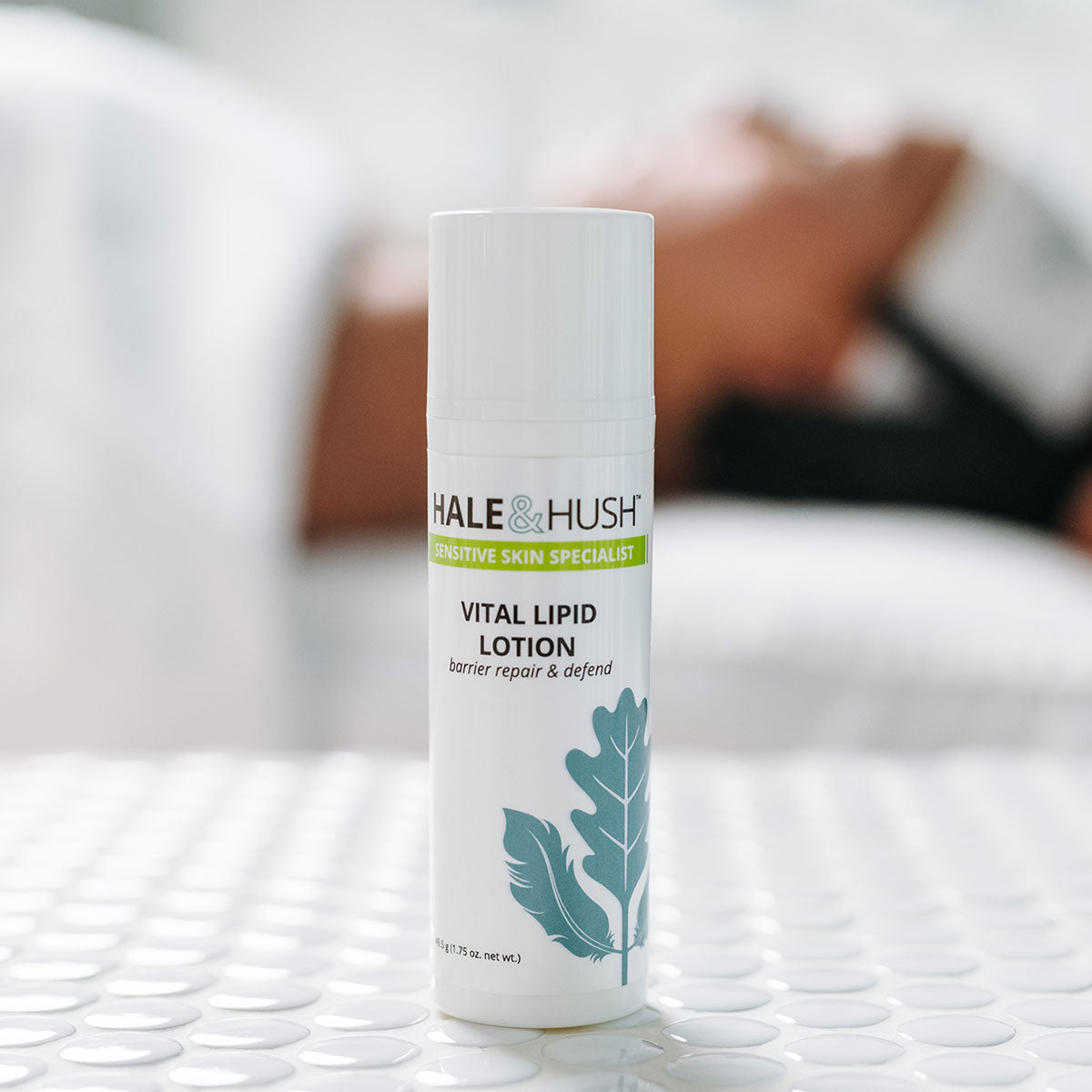 Hale & Hush's Vital Lipid Lotion white product bottle standing on white spa bed with a model receiving a facial in the background