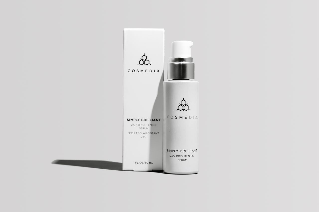 COSMEDIX's Simply Brilliant 24/7 Brightening Serum white colored product bottle with dispenser standing beside white colored product box, against light grey background