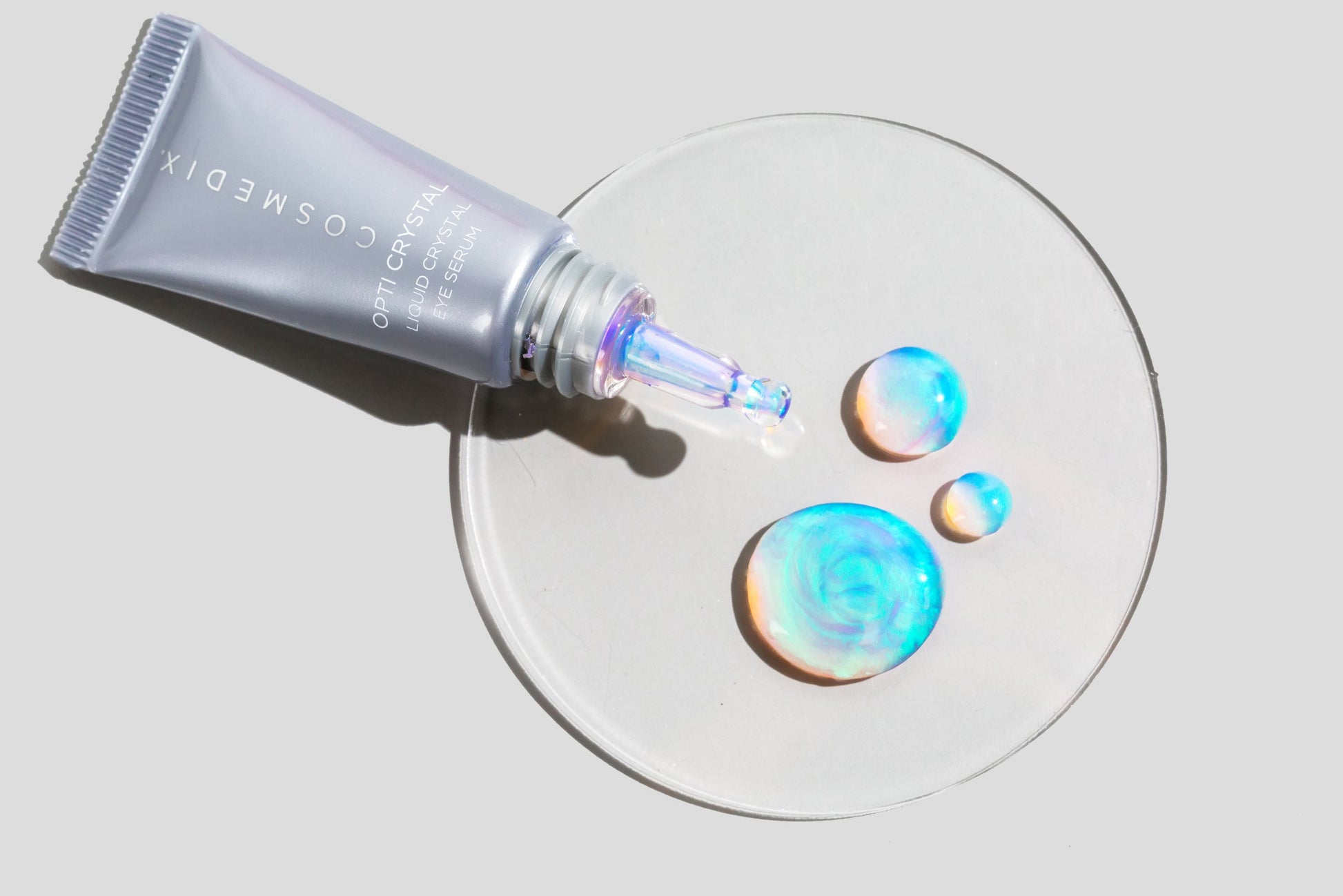 COSMEDIX's Opti Crystal Liquid Crystal Eye Serum light grey product bottle laying on its side with iridescent product swatches on petri dish with a light grey background