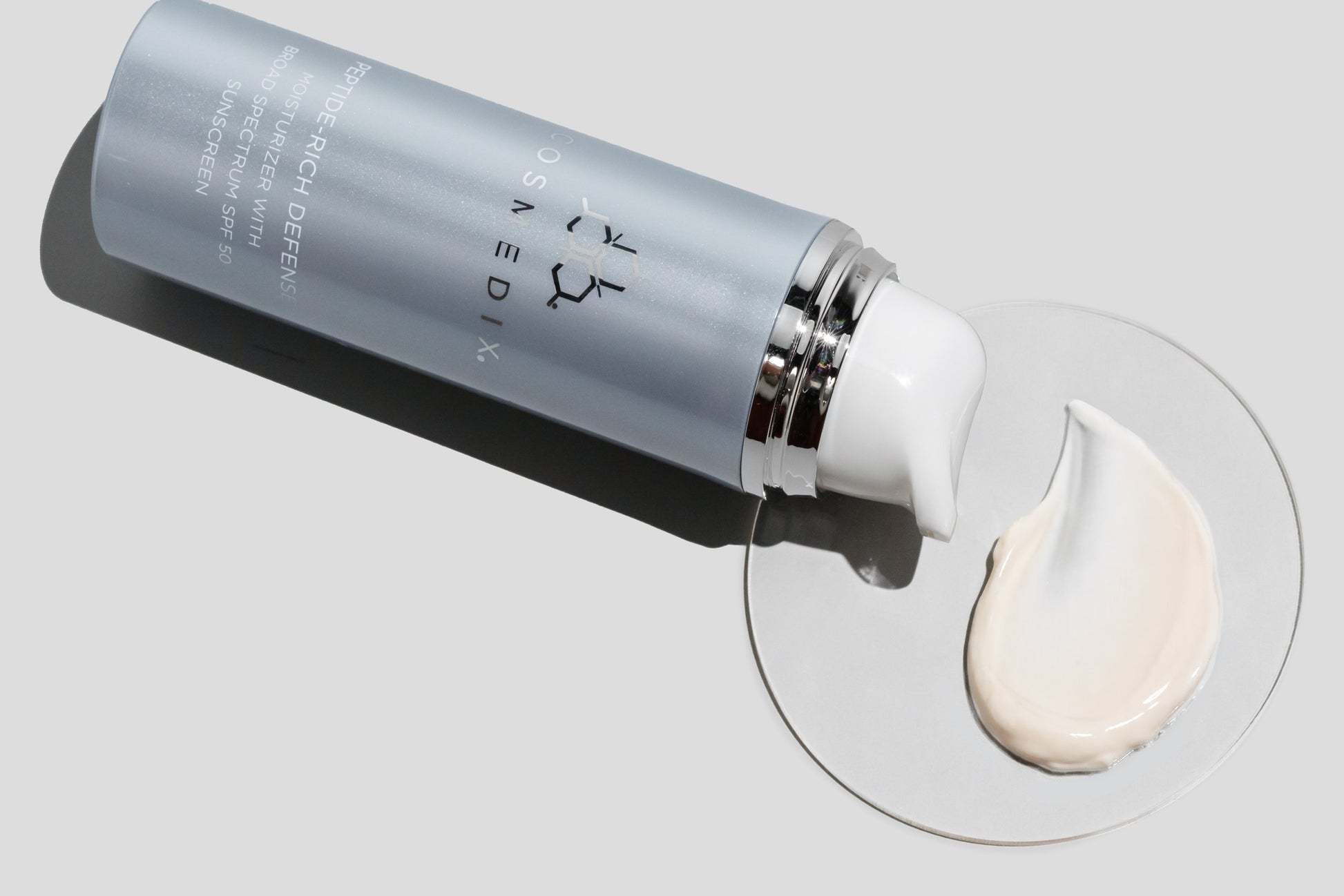 COSMEDIX'S Peptide-Rich Defense SPF 50 light grey product bottle laying on side with cream colored product swatch on a petri dish with a light grey background