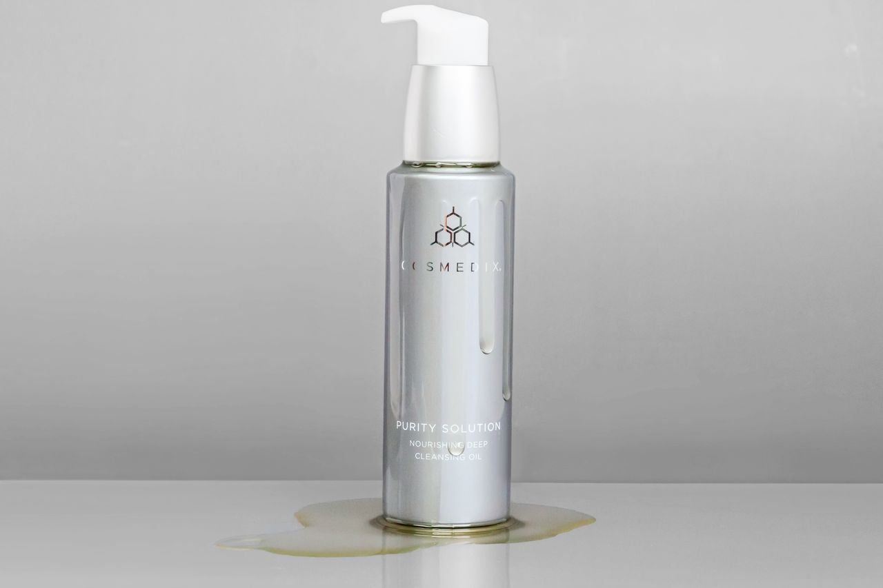 COSMEDIX's Purity Solution Nourishing Cleansing Oil light grey product bottle with white pump dispenser oozing gold colored product oil against grey background