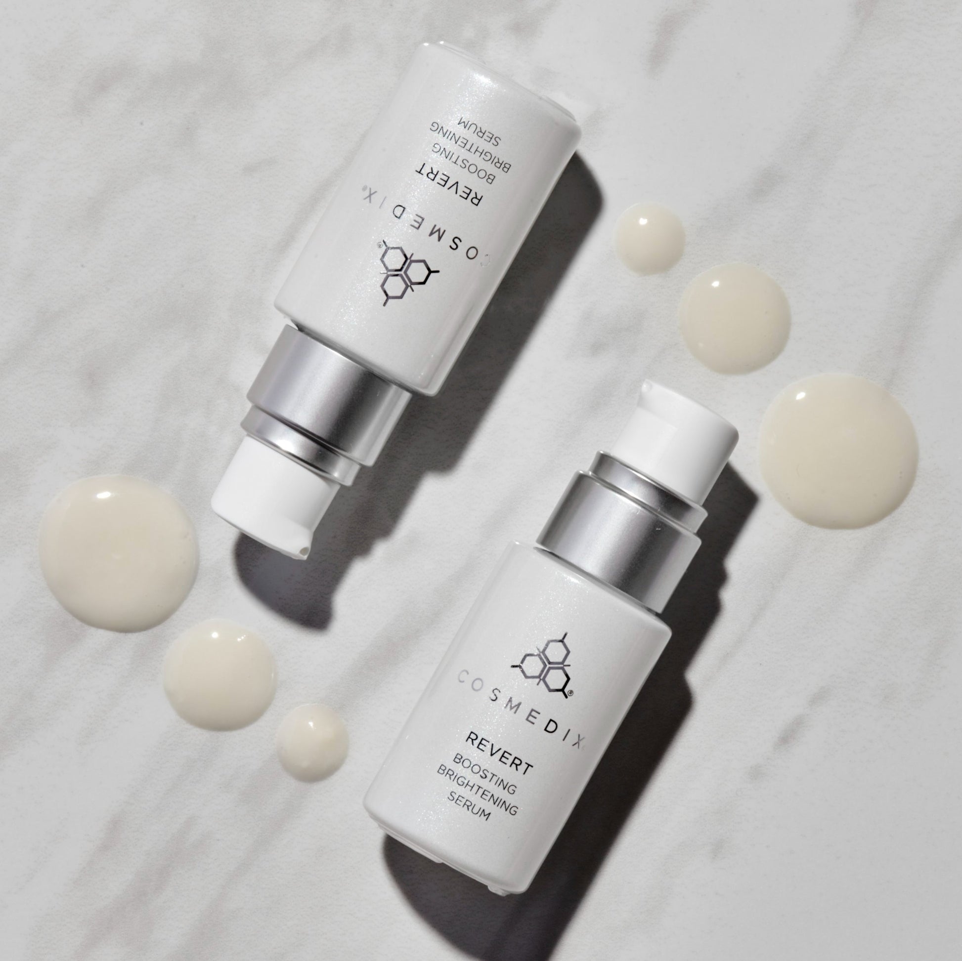 Two bottles of COSMEDIX's Revert Boosting Brightening Serum laying parallel with cream colored product droplets against white and grey background