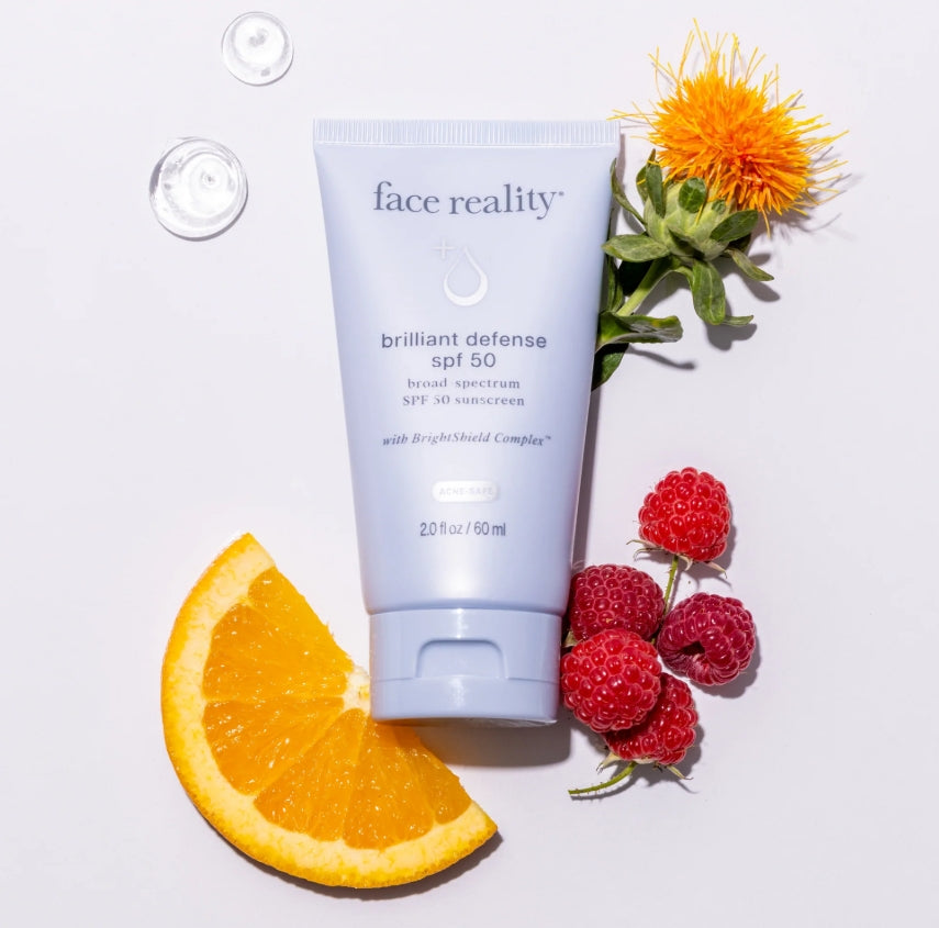 Face Reality's Brilliant Defense SPF 50 light blue product bottle lying beside an orange slice, raspberries, water droplets and an orange flower.