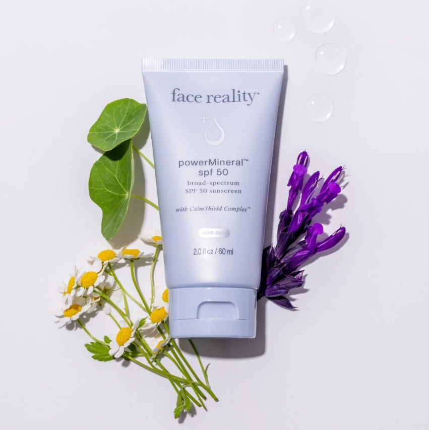 FaceReality's powerMineral SPF 50 tube lying beside assorted flowers and water droplets on an off-white background