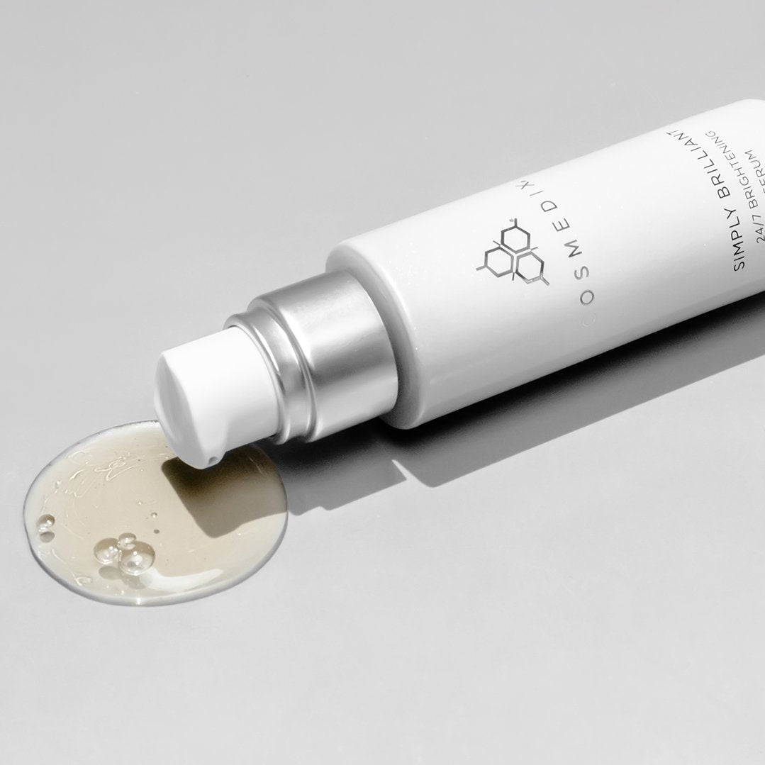 COSMEDIX's Simply Brilliant 24/7 Brightening Serum white product bottle laying on side with white dispenser and opaque colored product droplet against light grey background