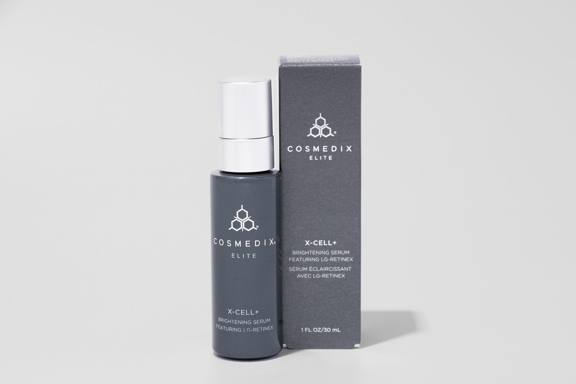COSMEDIX's X-Cell+ Brightening Serum dark grey product bottle with silver cap standing beside dark grey product box against light grey background