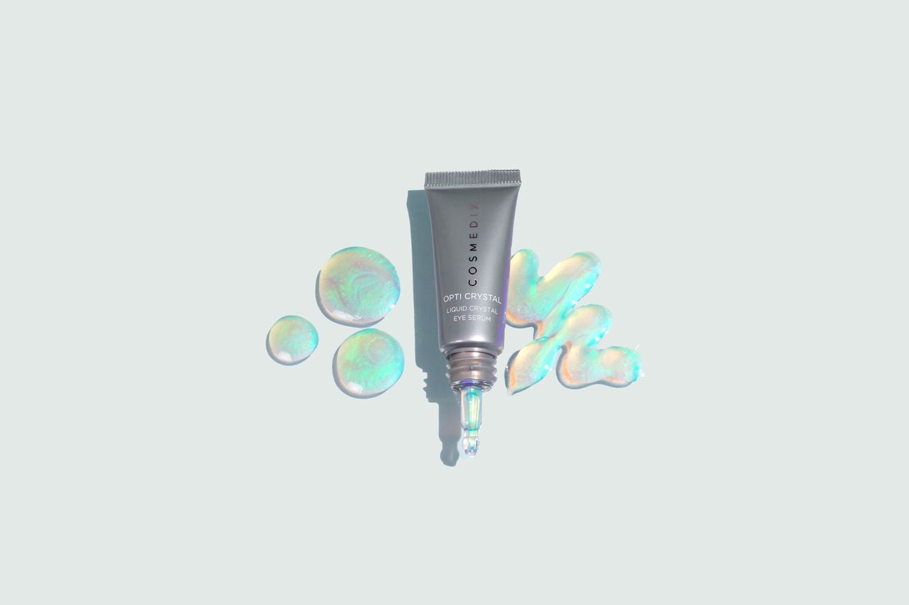 COSMEDIX's Opti Crystal Liquid Crystal Eye Serum grey product bottle with cap off, lying beside iridescent colored product swatches on a light grey background