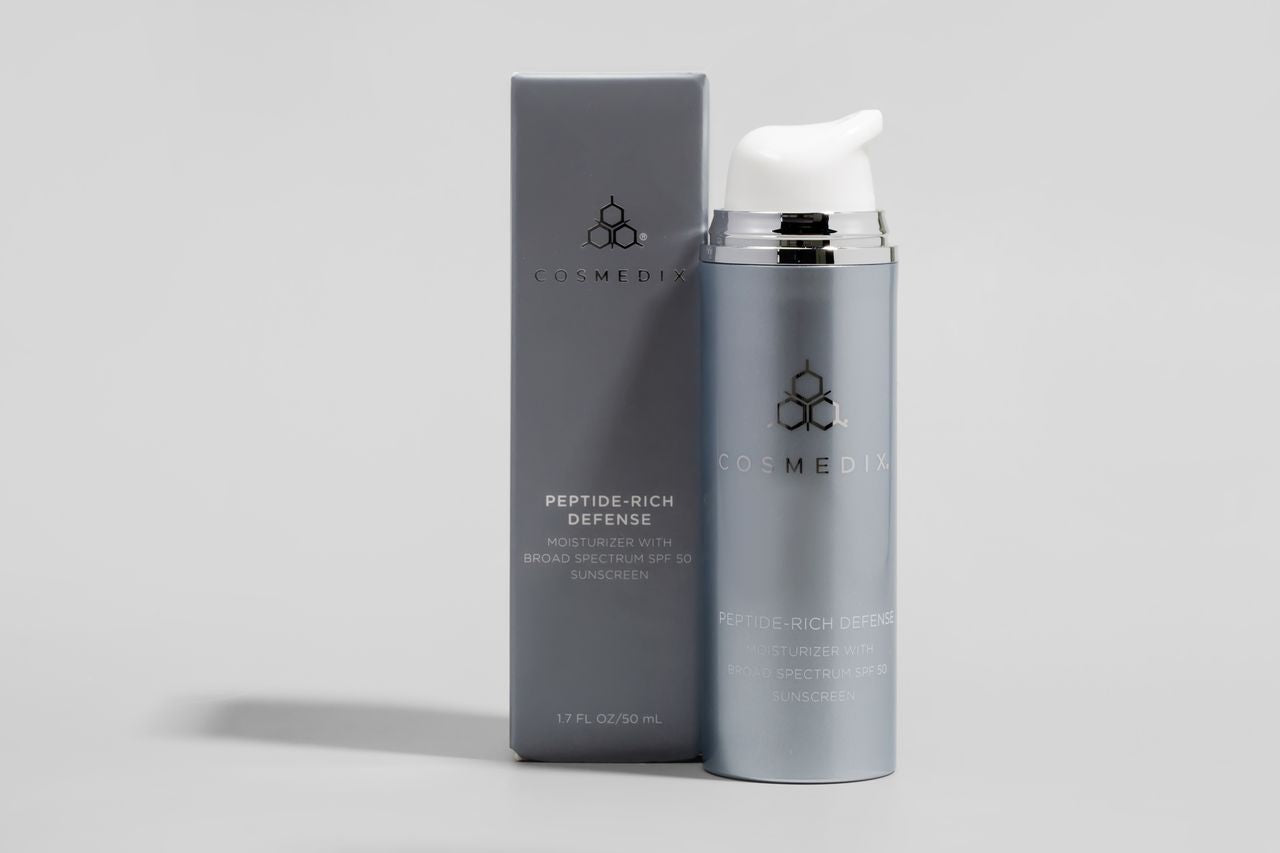 COSMEDIX'S Peptide-Rich Defense SPF 50 grey colored product bottle with white pump dispenser standing beside grey product box, against a light grey background