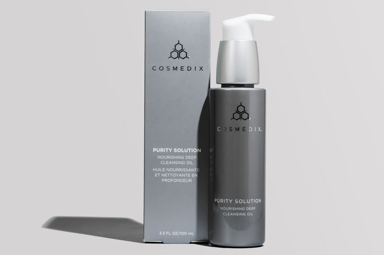 COSMEDIX's Purity Solution Nourishing Cleansing Oil grey product bottle with white dispenser standing beside light grey product box, against light grey background