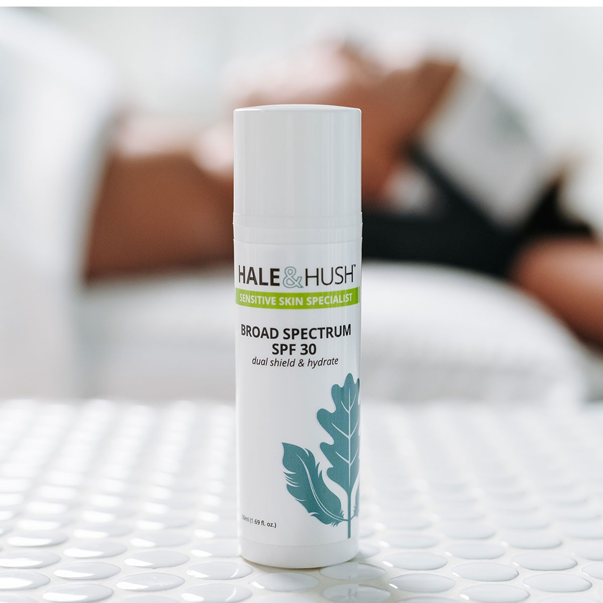 Hale & Hush's Broad Spectrum SPF 30 white product bottle standing on white spa bed with a model receiving a facial in the background