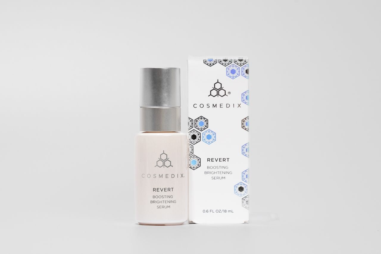 COSMEDIX's Revert Boosting Brightening Serum white product bottle with silver lid standing beside white product box with blue hexagons, against a light grey background
