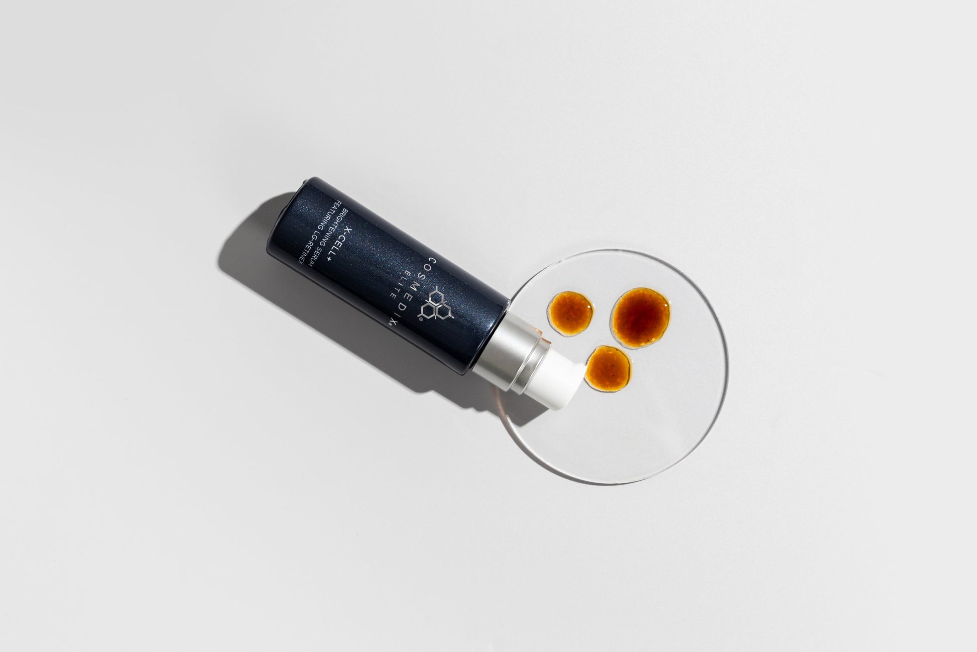 COSMEDIX's X-Cell+ Brightening Serum dark grey product bottle laying on side with dark orange product droplets on a petri dish against a light grey background